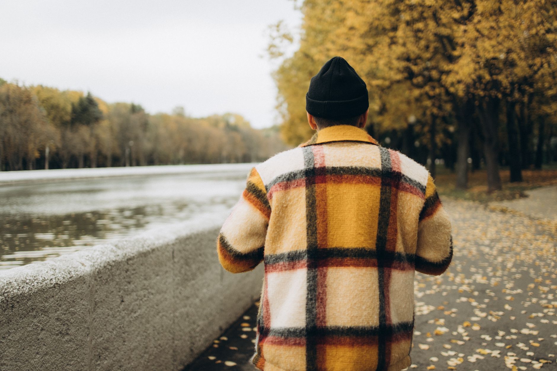 A person wearing a plaid jacket walks alongside a river in an autumn park.