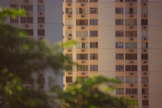 High-rise building facade with windows and air conditioners in focus, blurred trees in foreground.