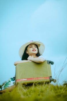 Asian woman in a sun hat leaning on luggage outdoors, smiling under blue sky.