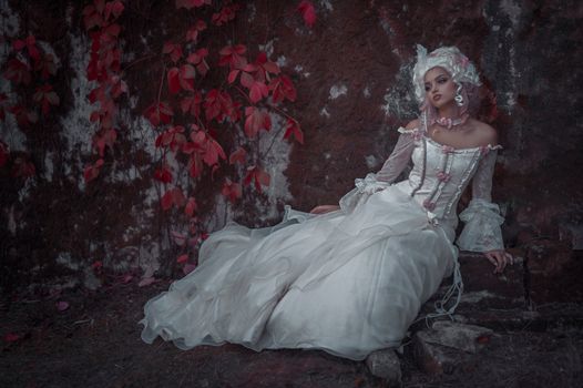 A woman in a vintage white dress poses against a rustic wall with red leaves, creating a whimsical atmosphere.