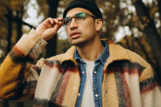 A fashionable man with eyewear and a checkered coat thoughtfully posing in a serene fall setting.
