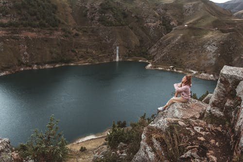 Free A Female Sitting at the Edge of a Rock Cliff with a Mountain Lake  Stock Photo