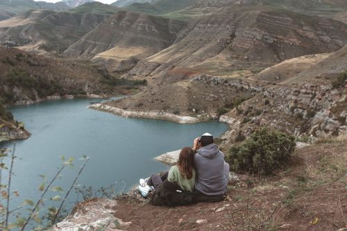 Free A couple sits atop a hill, capturing the stunning view of a tranquil mountain lake surrounded by hills. Stock Photo
