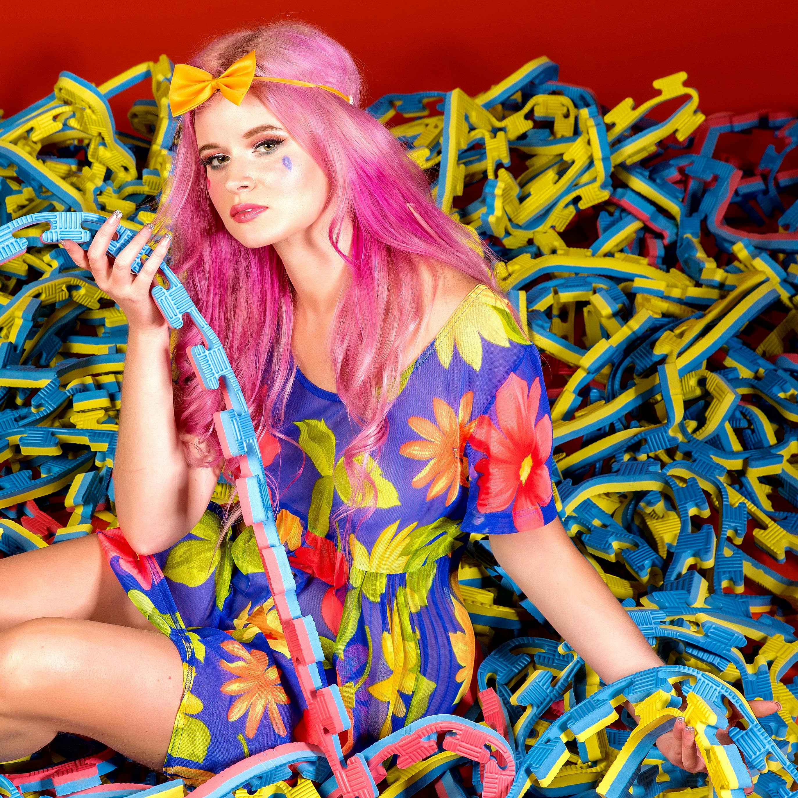 Fashion model with vibrant pink hair and floral dress surrounded by colorful toy pieces.