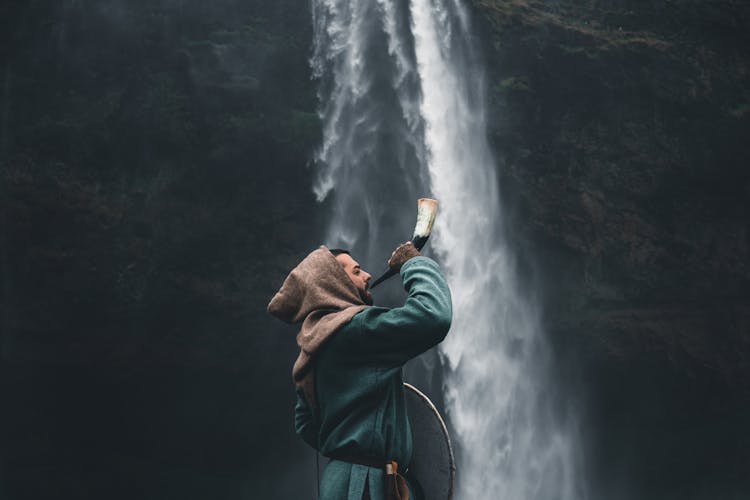 Man In Warrior Clothes Blowing Horn By Waterfall