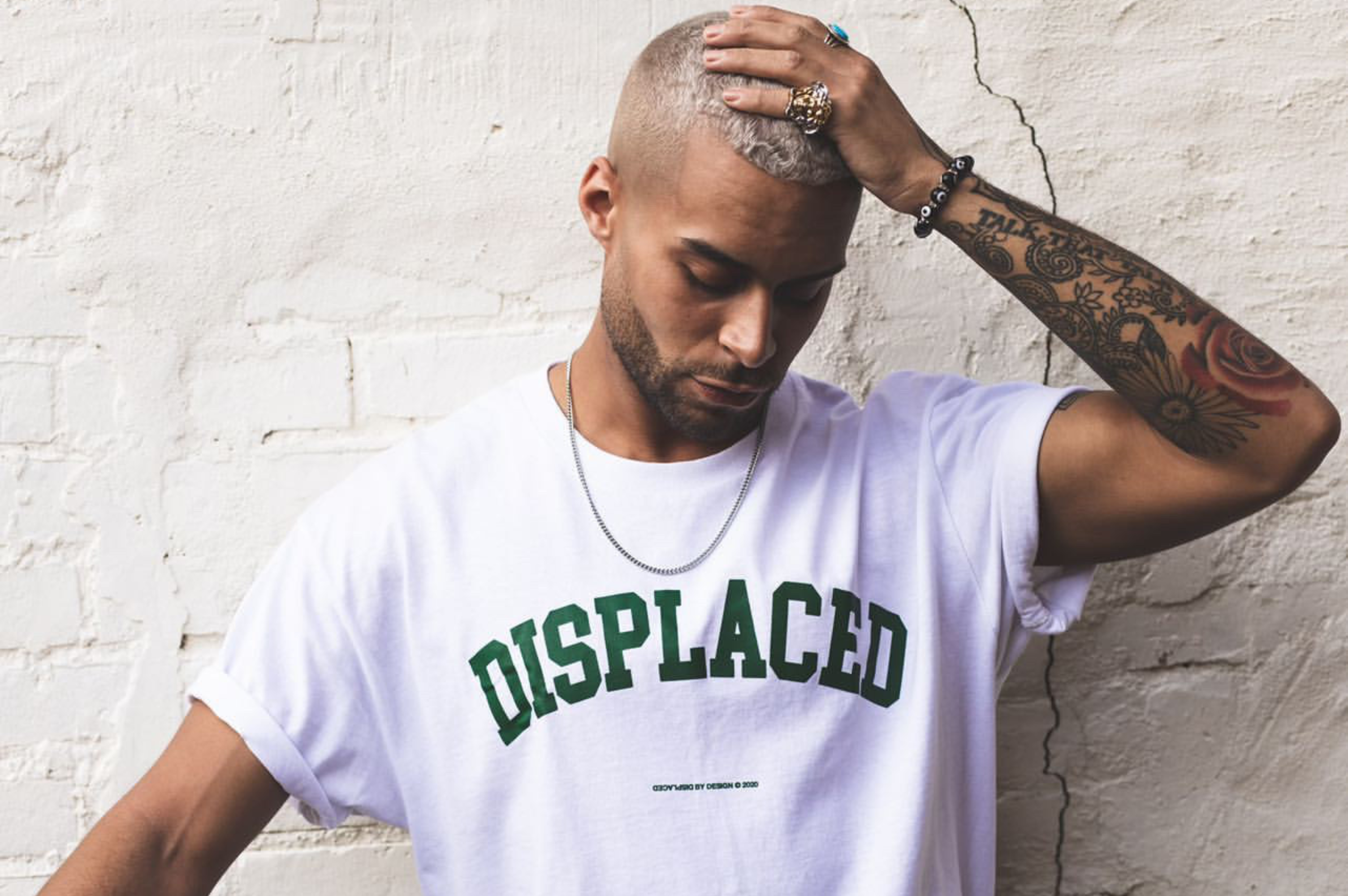 Man with arm tattoo wearing a 'DISPLACED' tee, posing thoughtfully indoors.