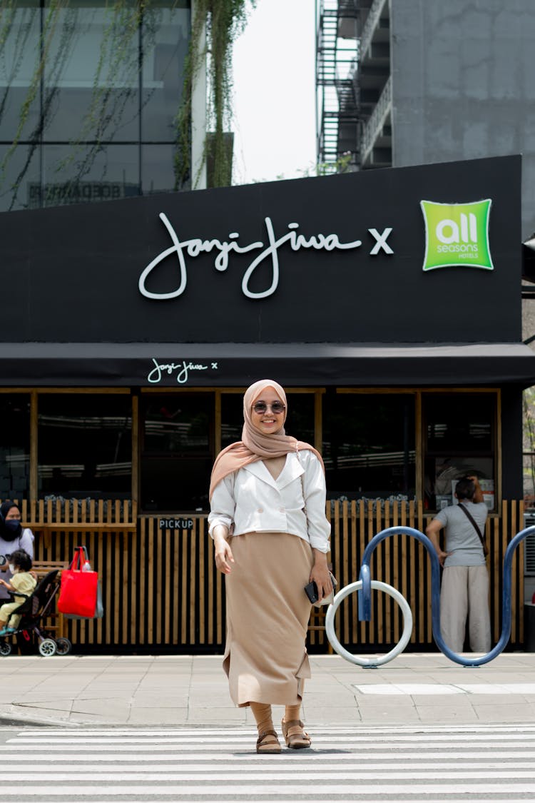 Woman Wearing Beige Headscarf And Skirt Walking On Zebra Crossing And Bar In Background