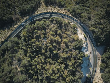 Scenic aerial view of a winding road through a lush forest with vehicles passing by.