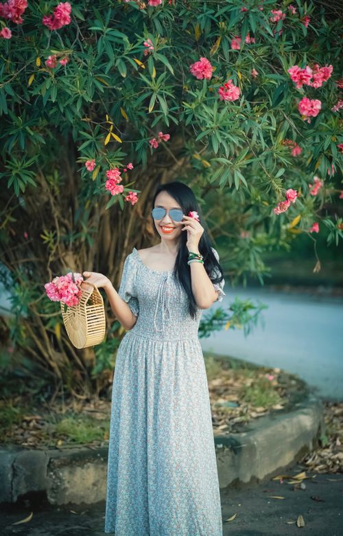 Free Chic woman in floral dress, holding bamboo handbag under blooming pink flowers outdoor. Stock Photo