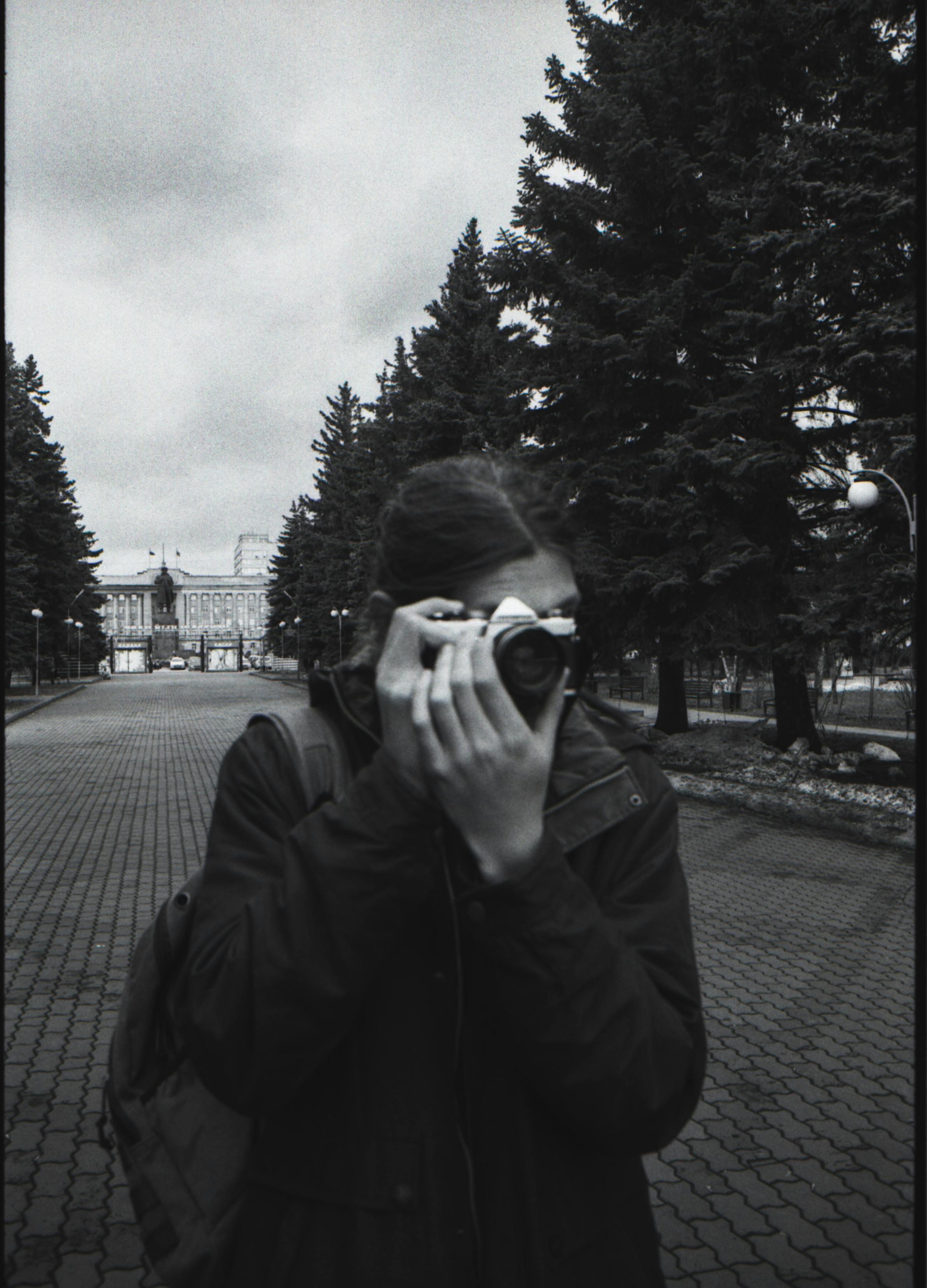 Person in black jacket using a rangefinder camera in a park setting.