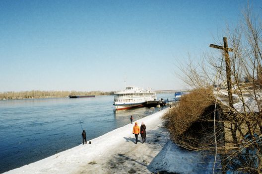 People walking along a snowy riverbank with a ship docked nearby under a clear winter sky.