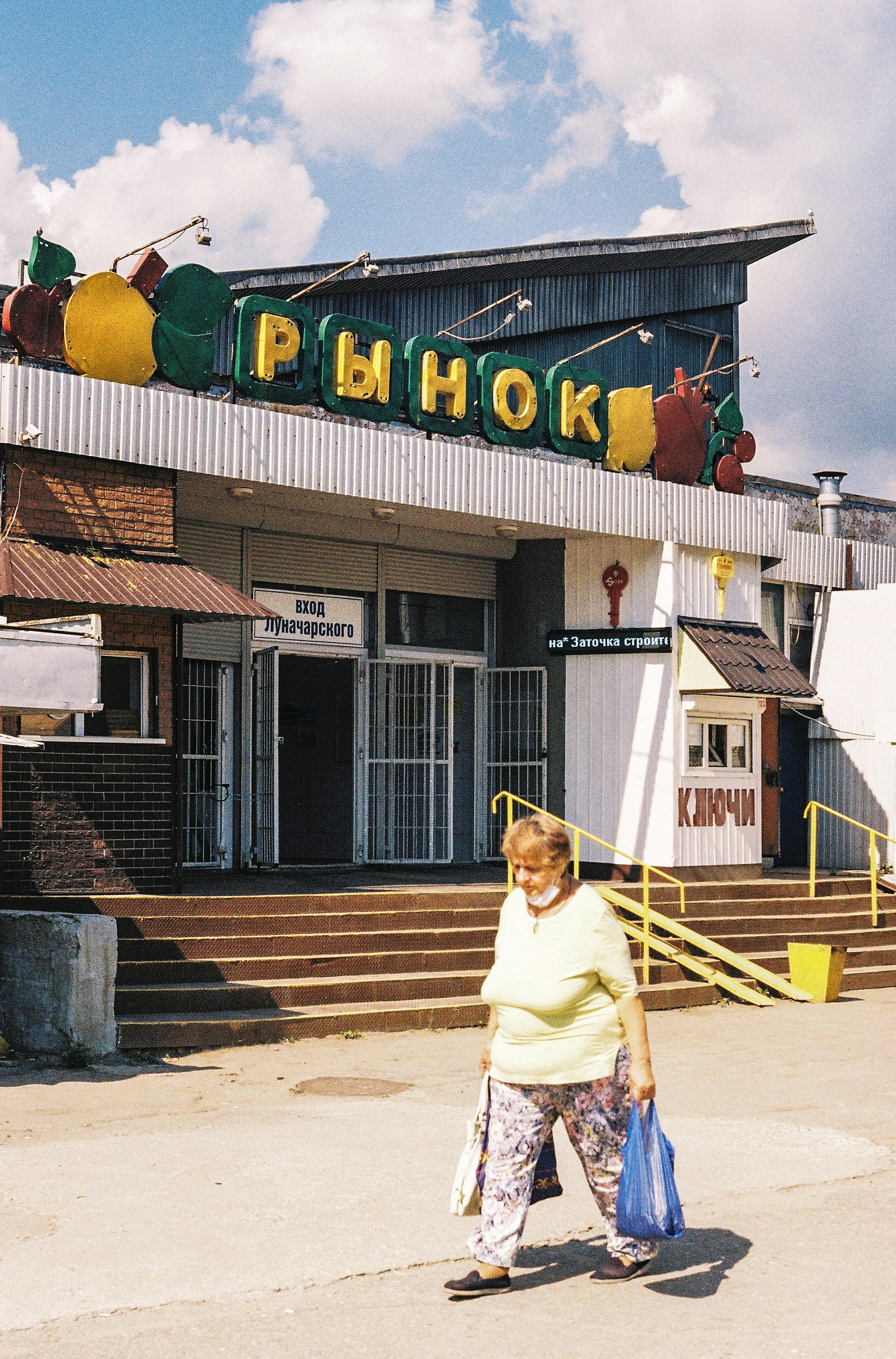 Elderly woman walking with groceries in front of outdoor market, sunny day.