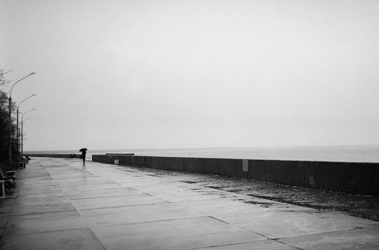 A solitary figure walks with an umbrella on a rainy seaside promenade.