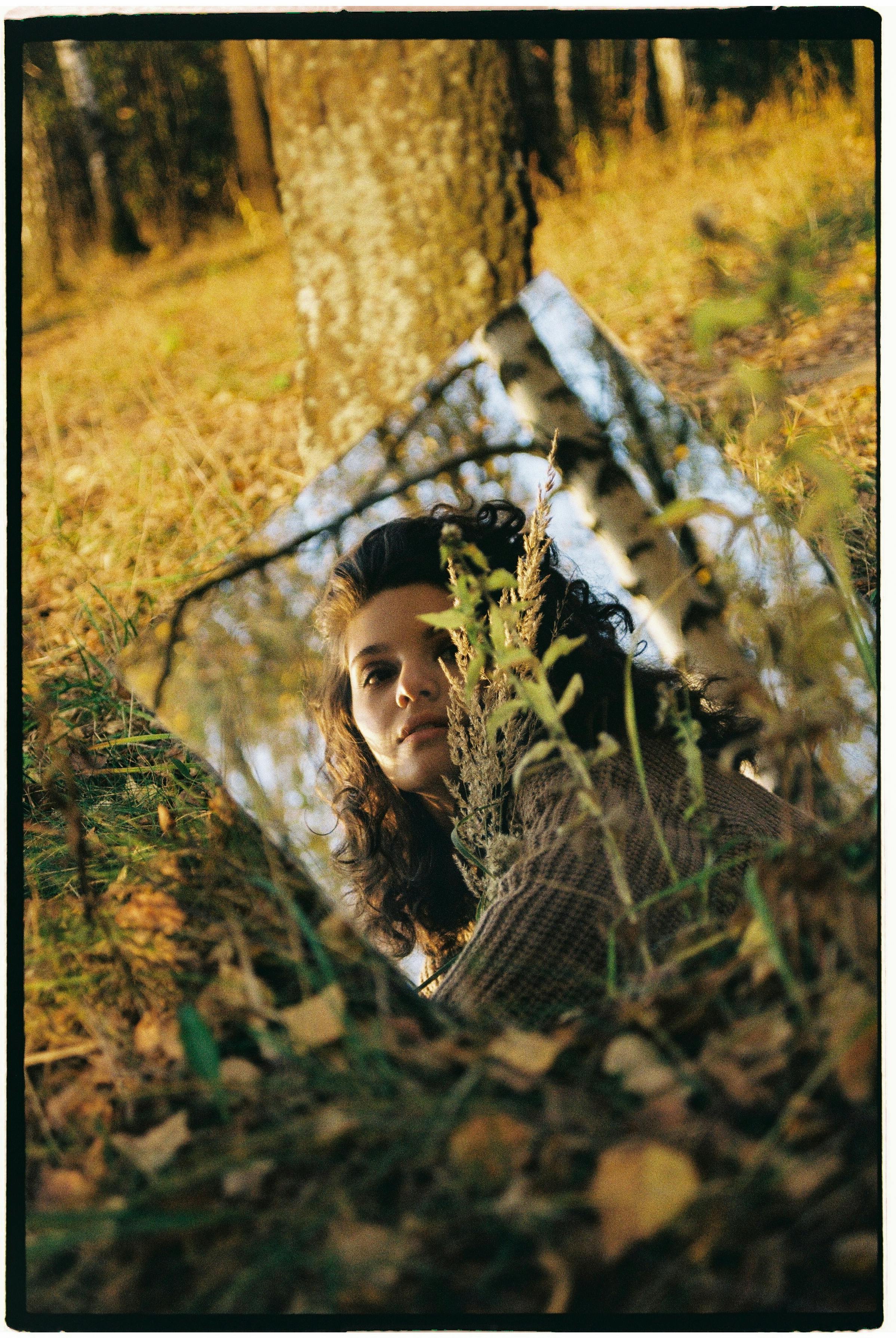 Artistic portrait of a woman reflected in a mirror, surrounded by fall foliage in a forest.