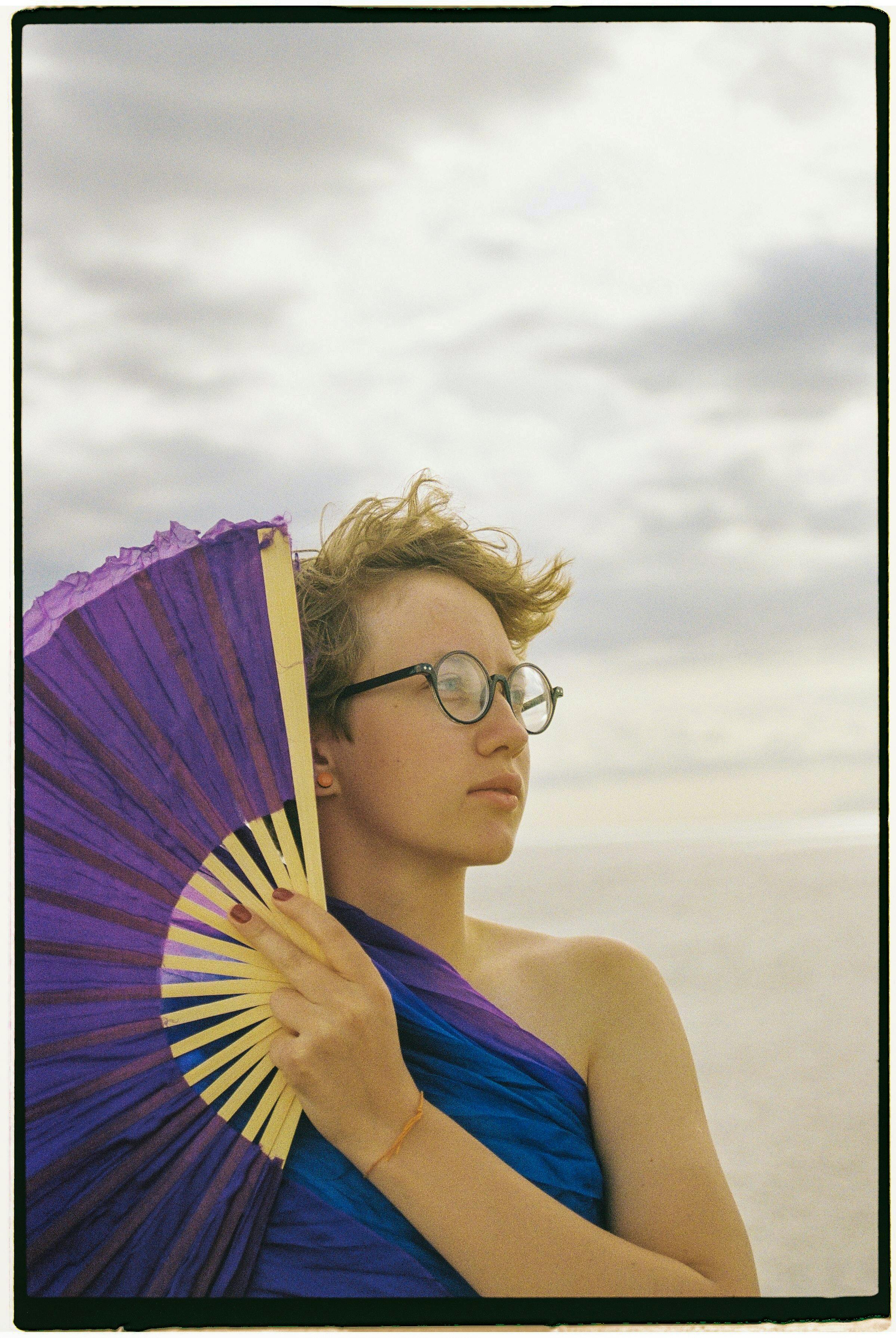 A woman stands on a beach holding a purple fan, looking thoughtful under a cloudy sky.