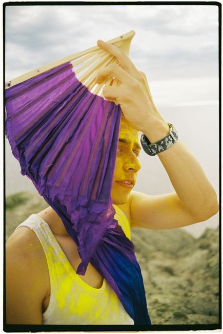 Colorful portrait of a person holding a violet fan outdoors, painted in yellow powder.