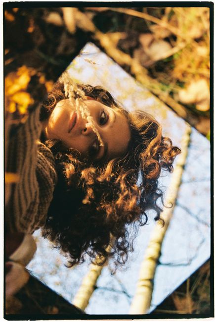 Young woman reflected in a mirror amidst autumn leaves, capturing a unique artistic portrait.