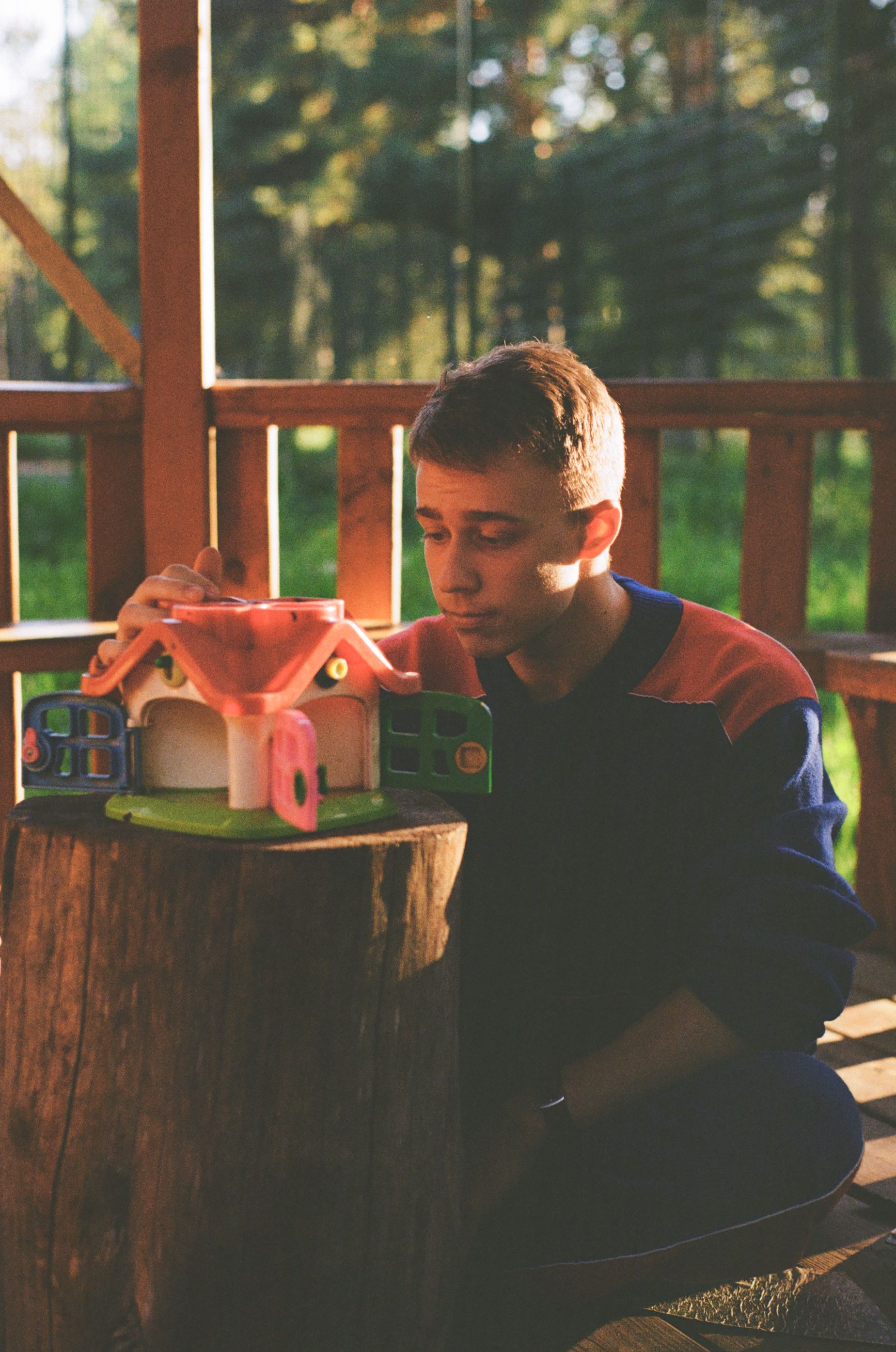 A man sits outdoors at sunset, focused on a toy house placed on a tree stump.