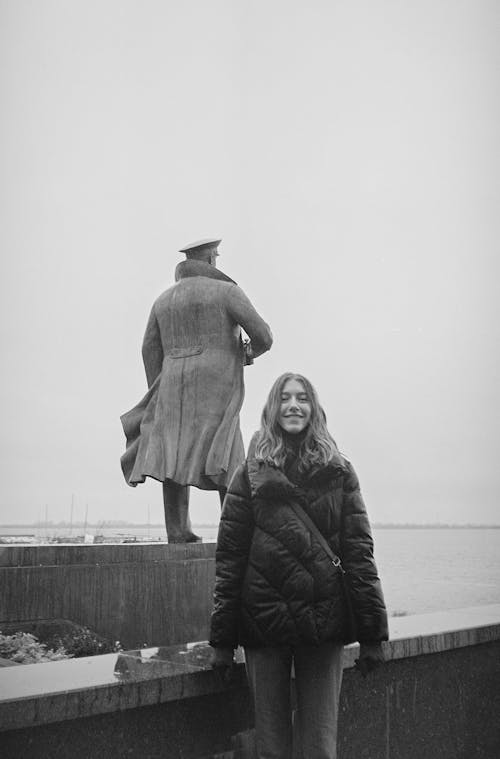 Black and White Photo of Woman in Black Jacket Standing near a Statue