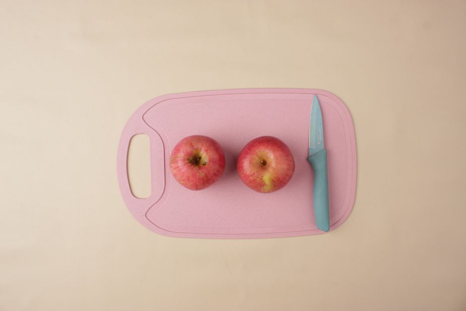Two fresh apples with kitchen knife on pink chopping board, beige background. Perfect for healthy lifestyle concepts.