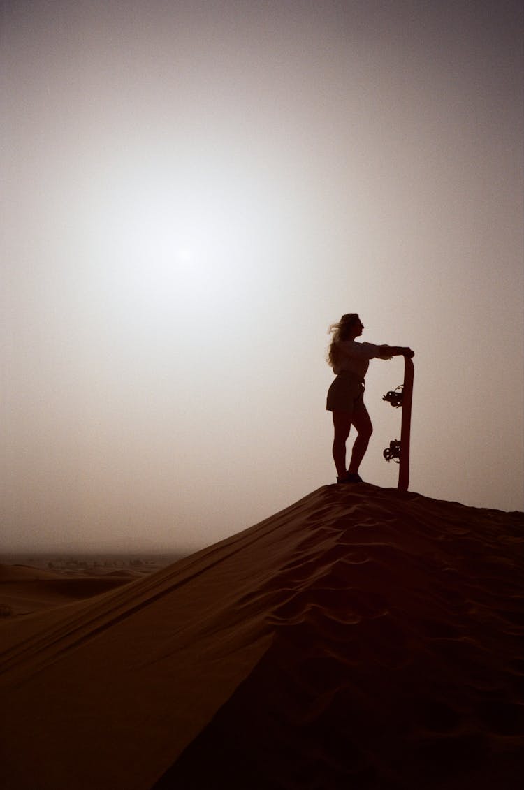 Woman Standing On Sand Dune With Surf Board
