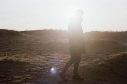 Silhouetted figure walking on a sandy beach with bright backlight from the sun.