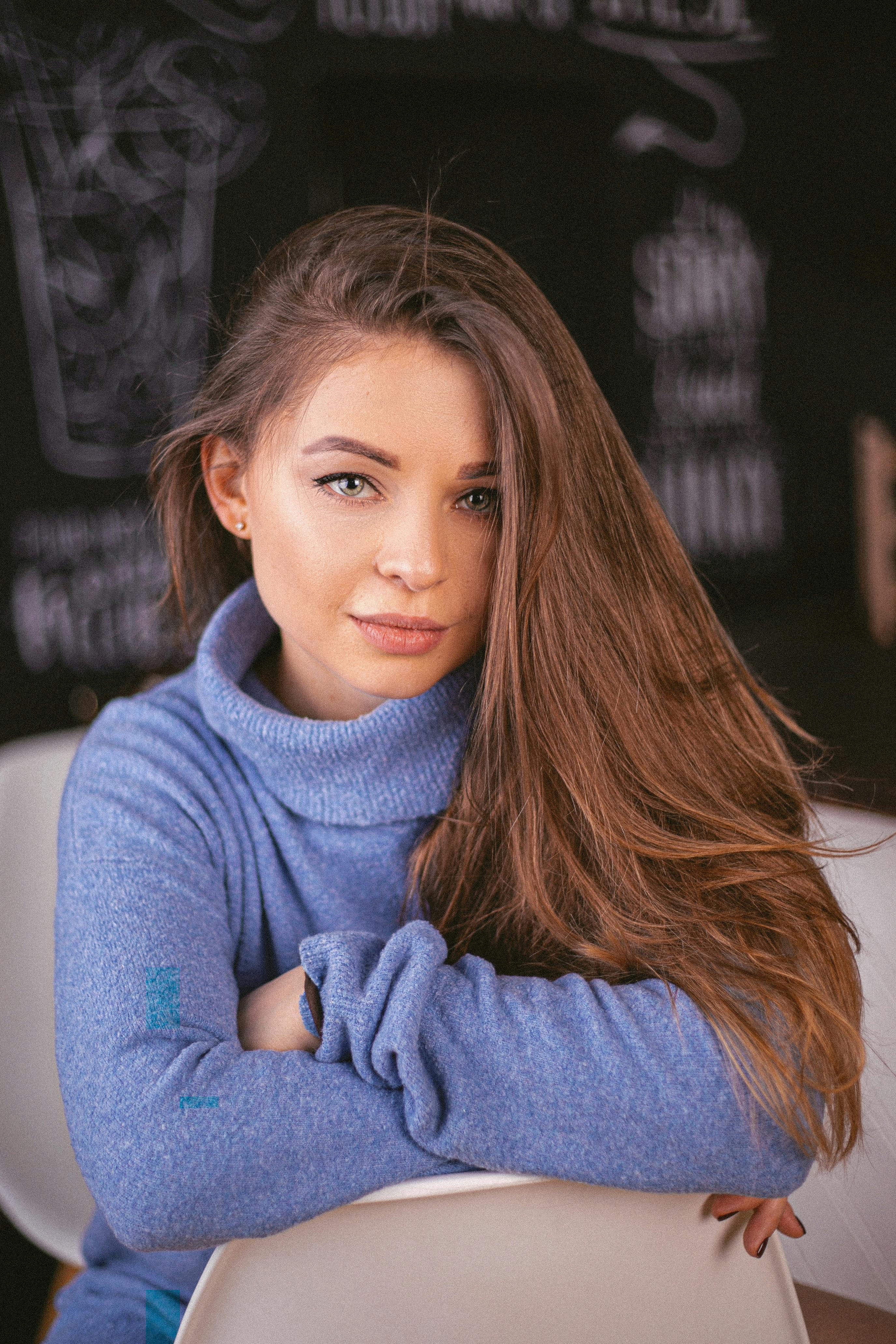 Portrait of a young woman with long brown hair wearing a blue sweater, seated indoors.