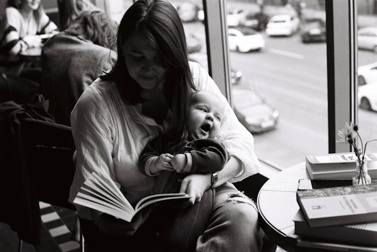 Woman In Long Sleeve Shirt Carrying A Baby While A Reading Book