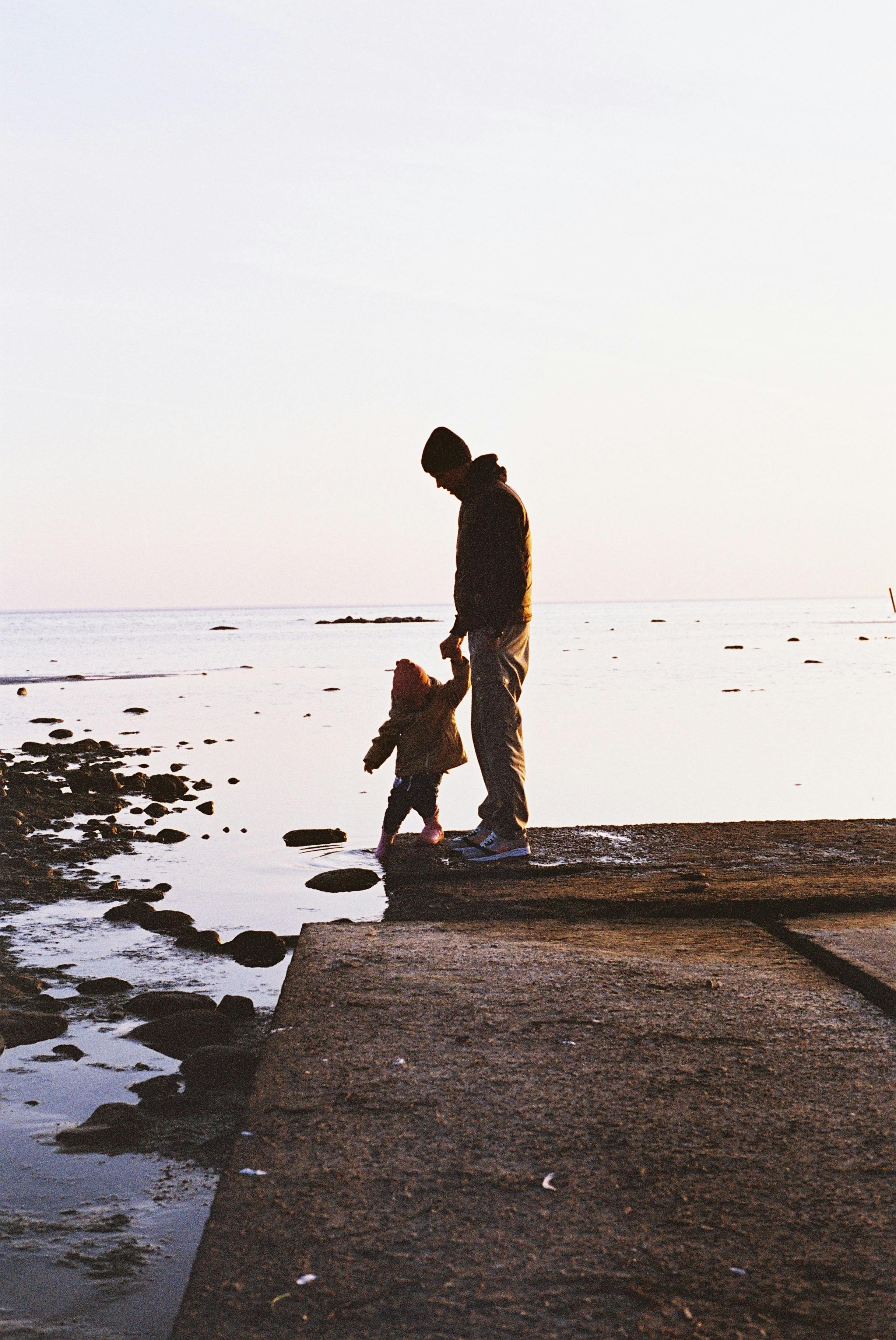 Tender moment of a father and child walking by the water's edge during sunrise. Perfect for themes of family and nature.