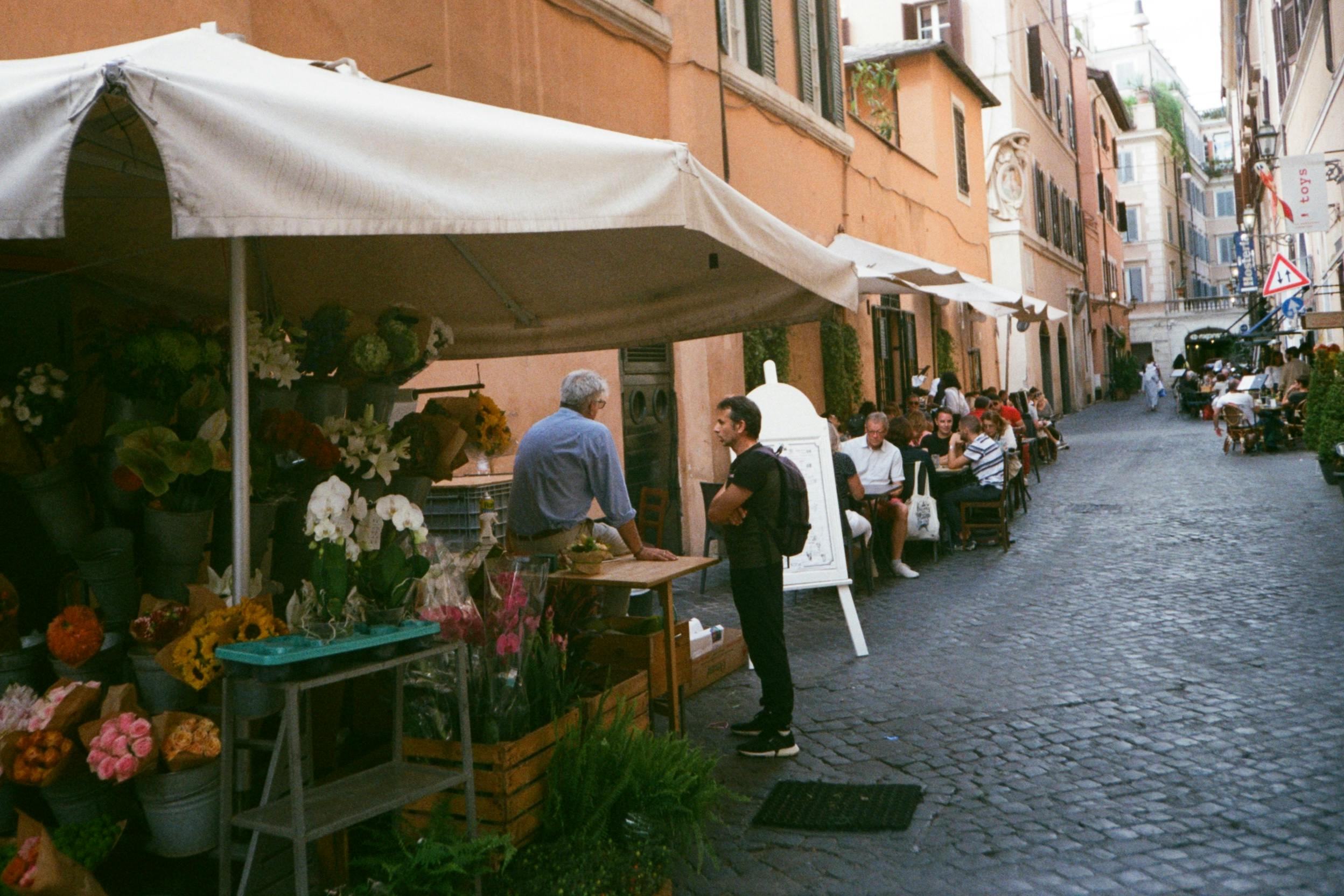 Explore a lively street scene in Rome featuring a flower stall and people enjoying outdoor cafes.