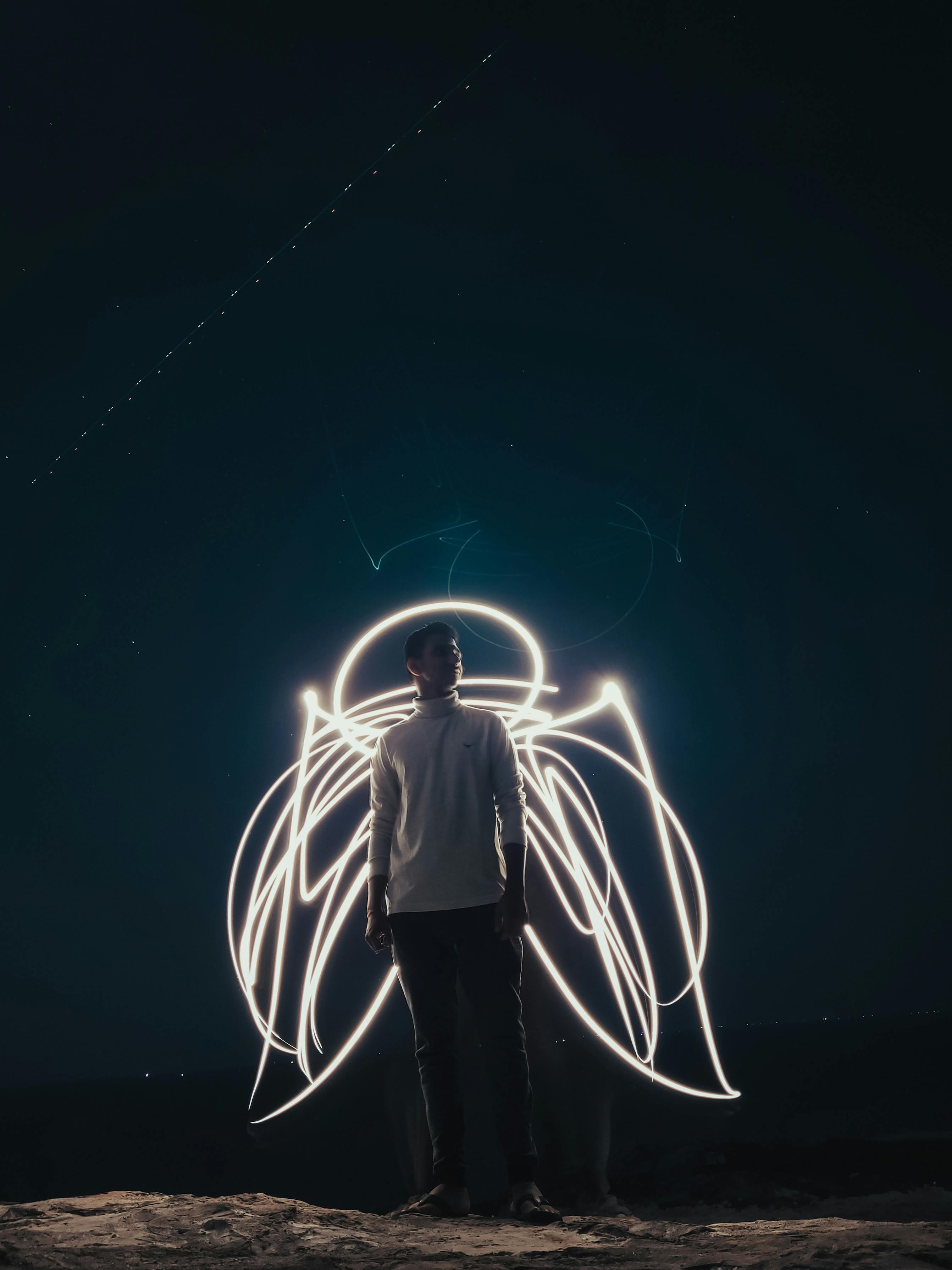 Silhouetted man surrounded by illuminated angel wing light trails under a night sky.