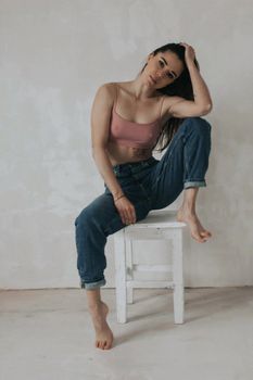 Young woman in jeans and bra posing on a stool indoors in a studio setting.