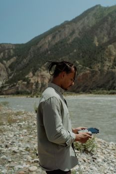 A man in a gray shirt operates a gadget by a riverside with mountains in the background.
