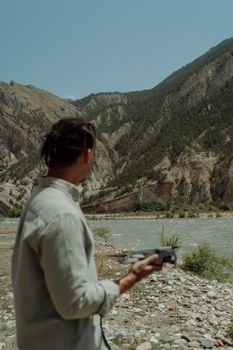 A man with dreadlocks holds a drone by a riverside, surrounded by mountains.