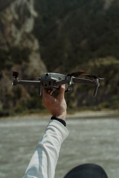 Person holding a drone by a river with mountains in the background, emphasizing outdoor technology.