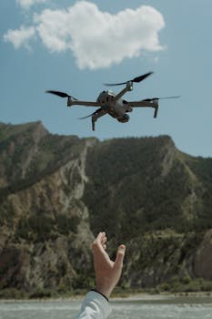 A drone flying against a mountainous backdrop with a person's hand reaching up, showcasing modern technology in nature.