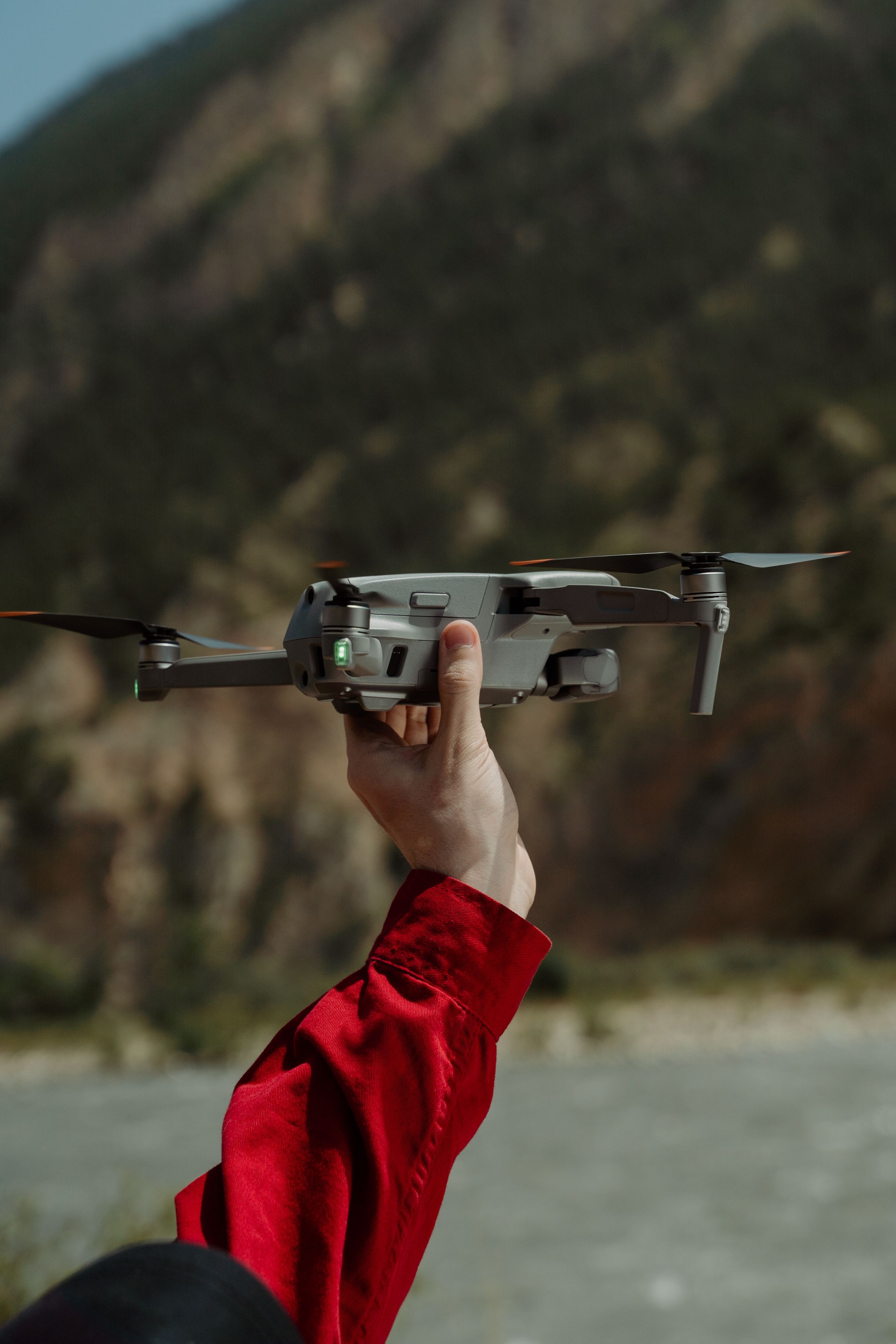 A person in a red jacket holding a drone outdoors in a mountainous area.