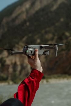 A person in a red jacket holding a drone outdoors in a mountainous area.