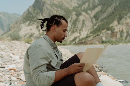 Free A man sits by a river, reading a map amidst scenic mountain views, embodying adventure and exploration. Stock Photo