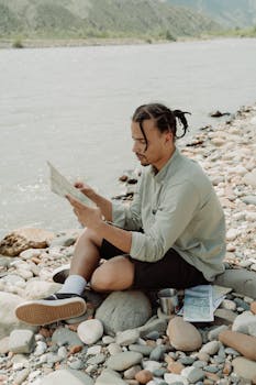 Man reading a map by a scenic riverside on a rocky bank.