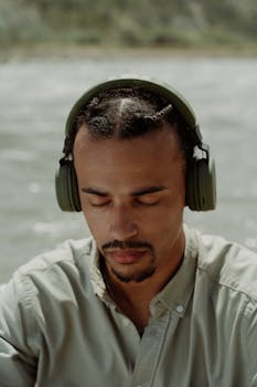 A man with headphones relaxes outdoors by a body of water, eyes closed, enjoying music.