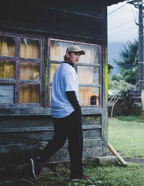 Man in White T-Shirt and Black Pants Walking by Windows of Wooden Cabin