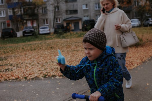 A young boy rides a scooter in a city street, capturing autumn's beauty with a phone call in the background.