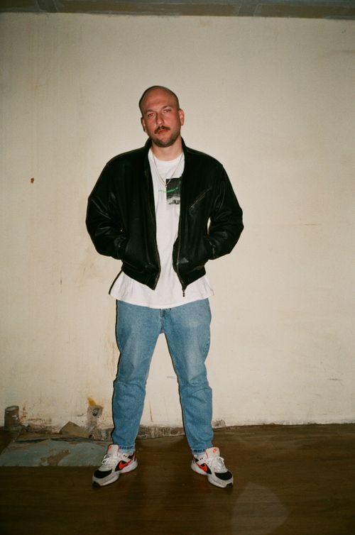 Free Adult man standing indoors in a black leather jacket and denim jeans against a white wall. Stock Photo