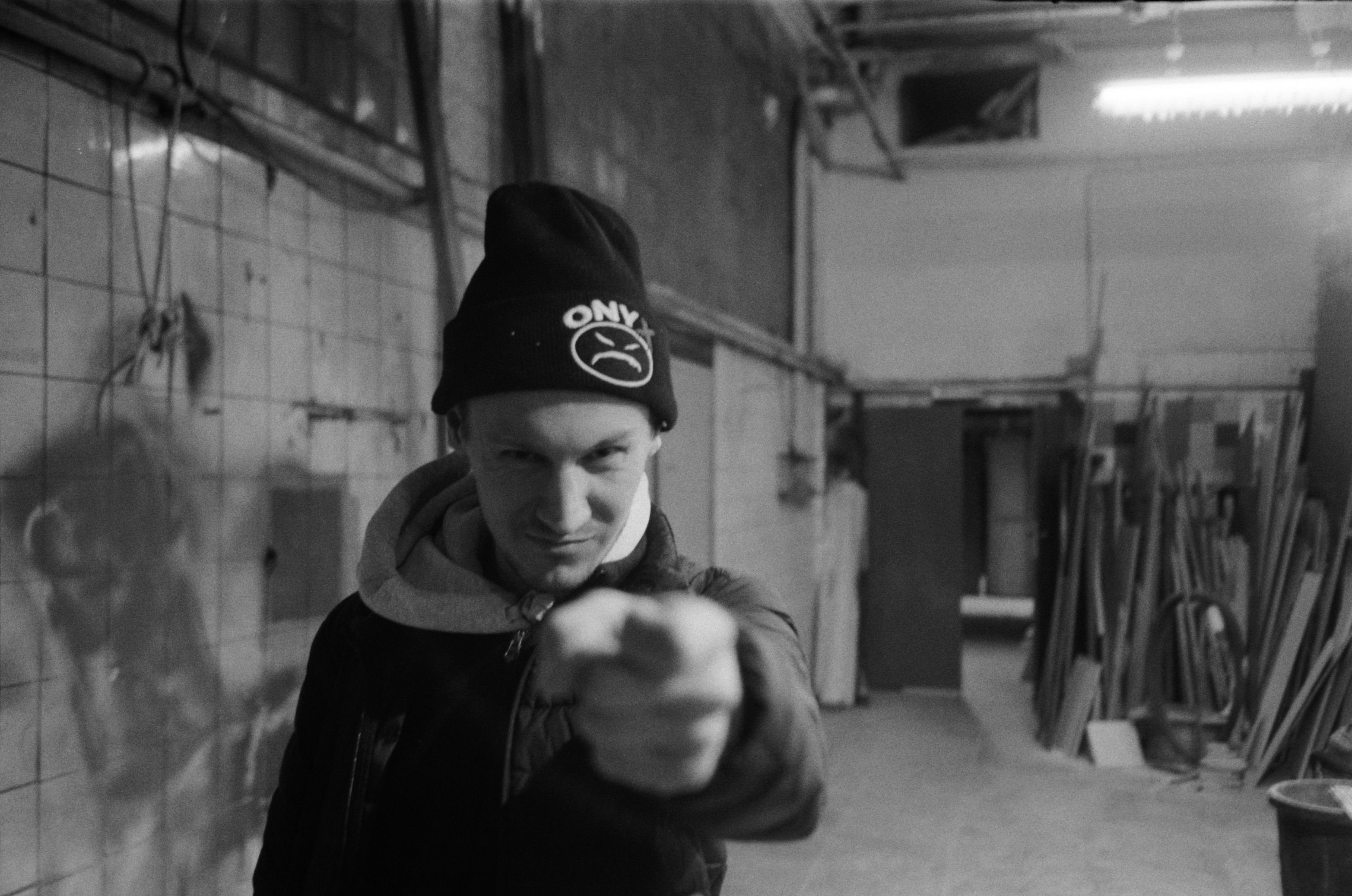 Grayscale image of a man in beanie and hoodie, pointing finger indoors.