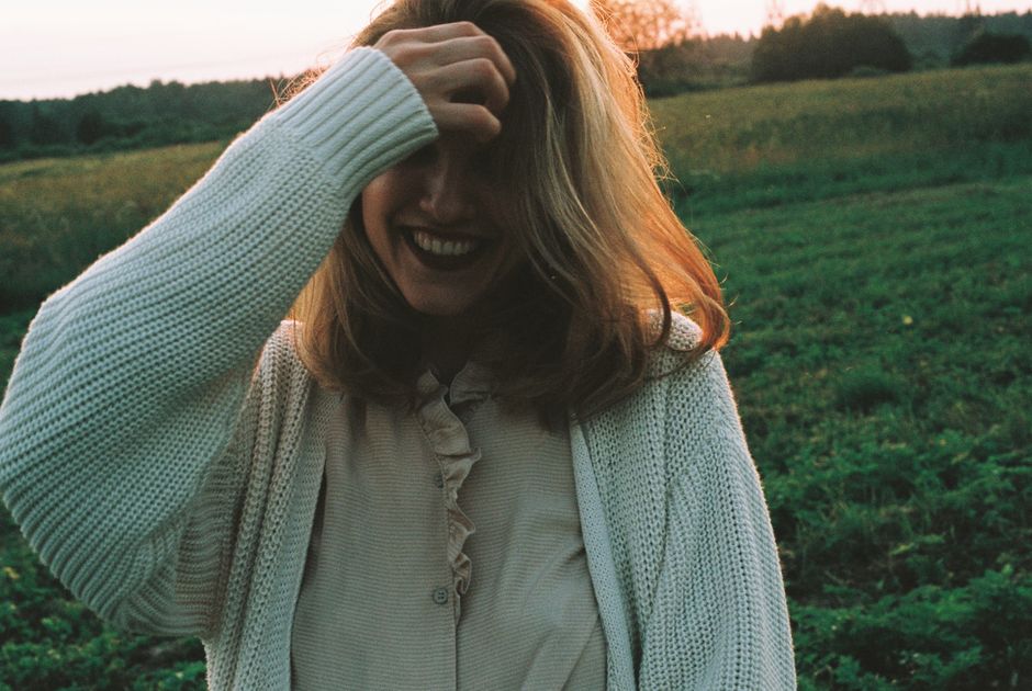 A cheerful woman with blonde hair smiles in a field at sunset, wearing a cozy knit sweater.