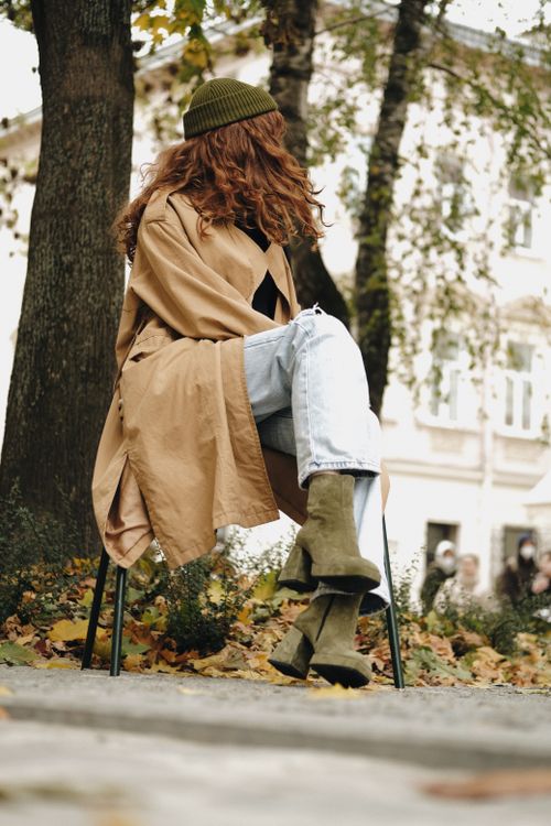 Free A stylish woman in a trench coat relaxes on a chair outdoors surrounded by autumn leaves. Stock Photo