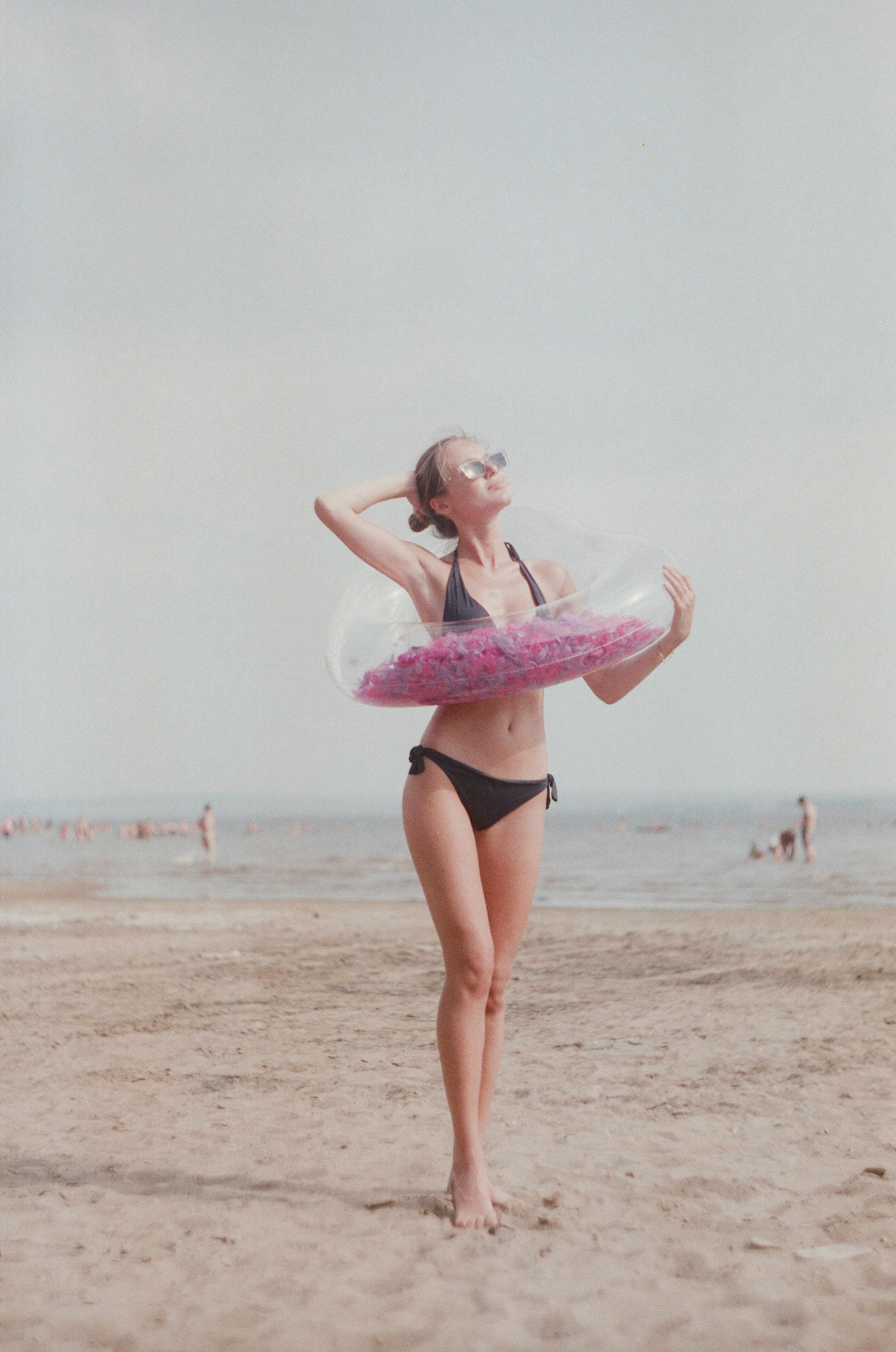 A woman in a bikini enjoys the beach with a pink floater under a clear sky.