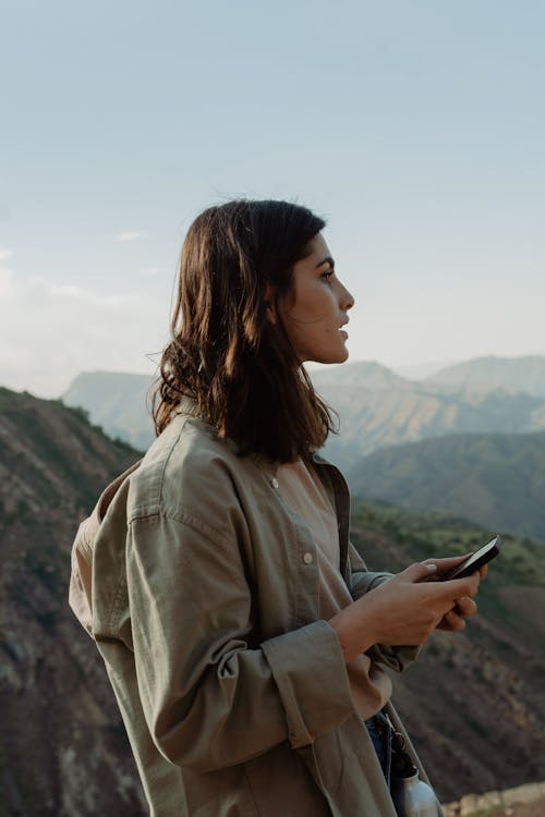 Woman Wearing Jacket and Holding Smartphone Standing Near Mountain View