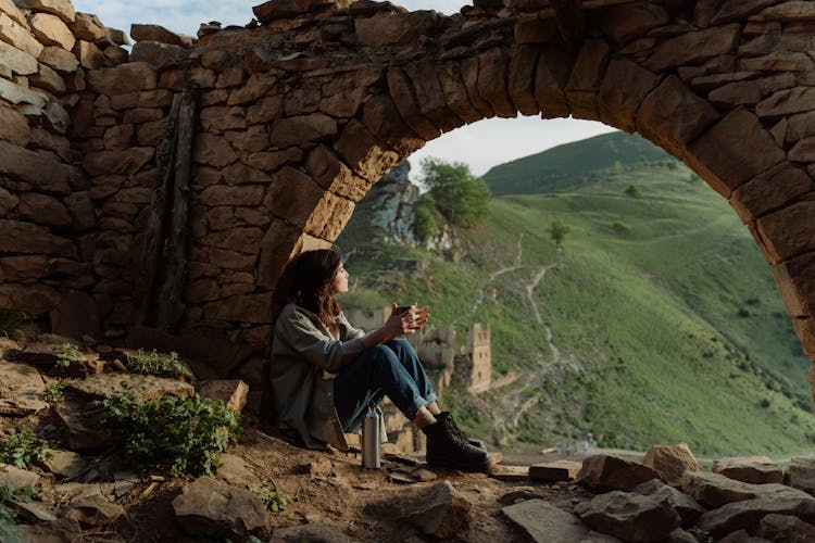 A Woman In Denim Jeans Sitting Under The Rock Formation While Looking At The View